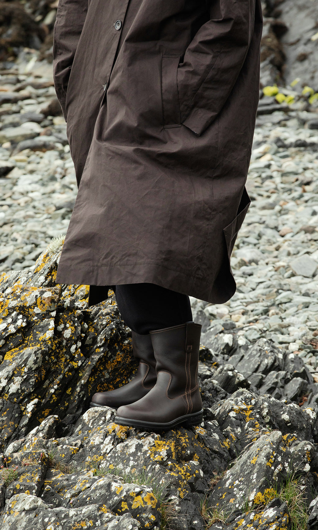 Plümo  Person in a brown coat and Rigger Boots stands on rocky ground covered with moss and lichen.