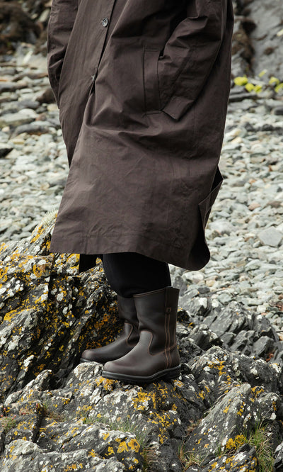 Plümo  Person in a brown coat and Rigger Boots stands on rocky ground covered with moss and lichen.