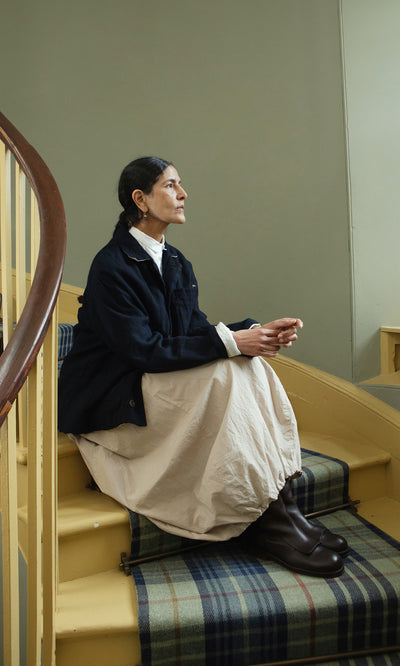 Plümo  Woman with dark braided hair sits on yellow stairs in the Dunbar Tweed Jacket and beige skirt, embodying slow fashion.