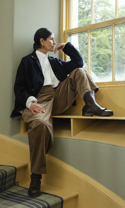 Plümo  Person in a Dunbar Tweed Jacket and brown pants sits on a windowsill, looking thoughtfully out the window.