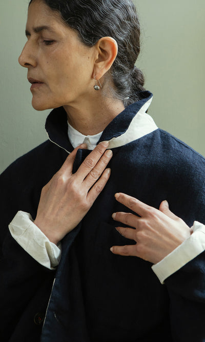 Plümo  Woman in a Dunbar Tweed Jacket adjusts her collar, looking down and embracing slow fashion against a pale backdrop.