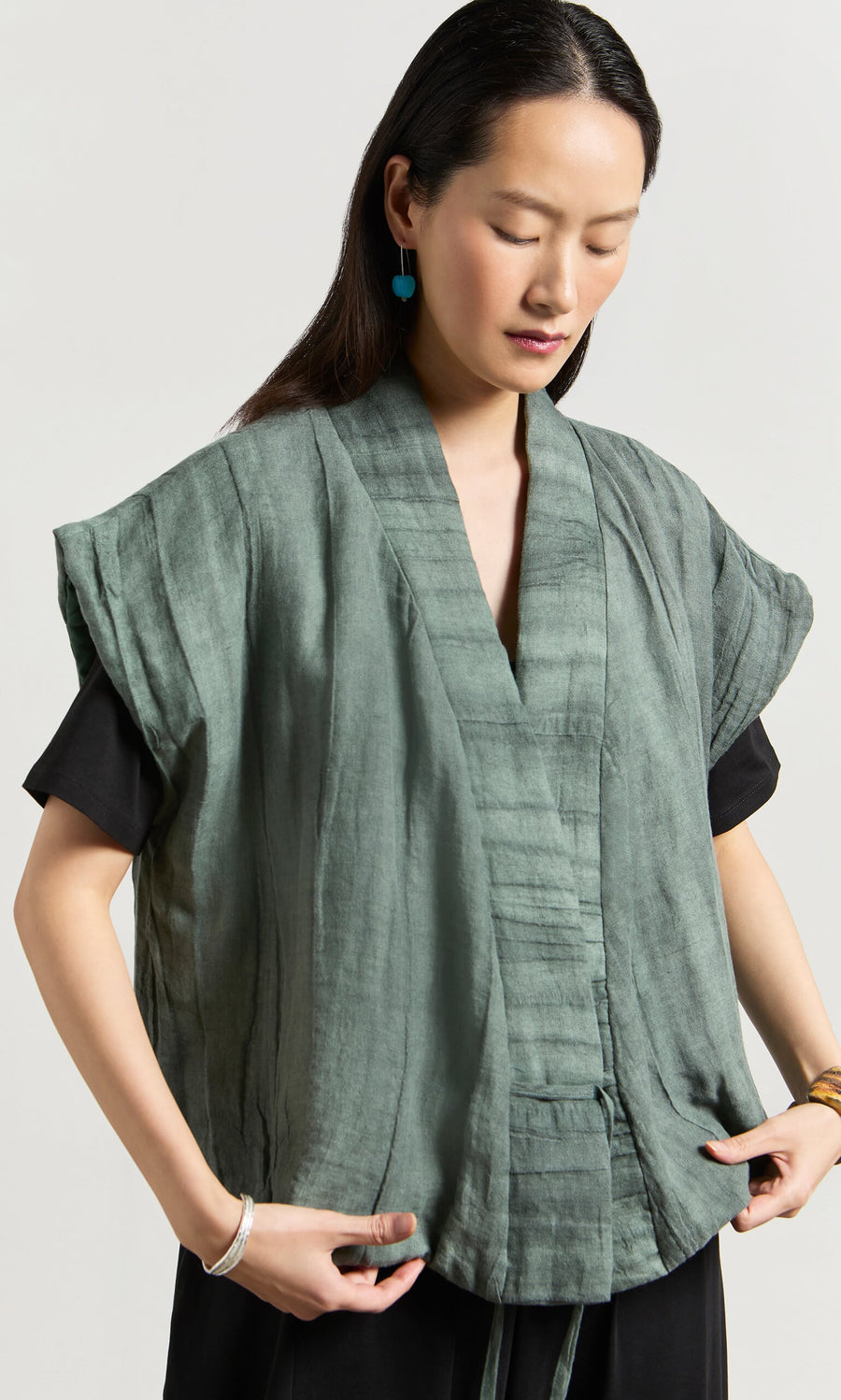 Plümo- Woman wearing the Fawn Gilet, a loose green textured wrap top, over a black shirt with blue earrings and gold bracelet.