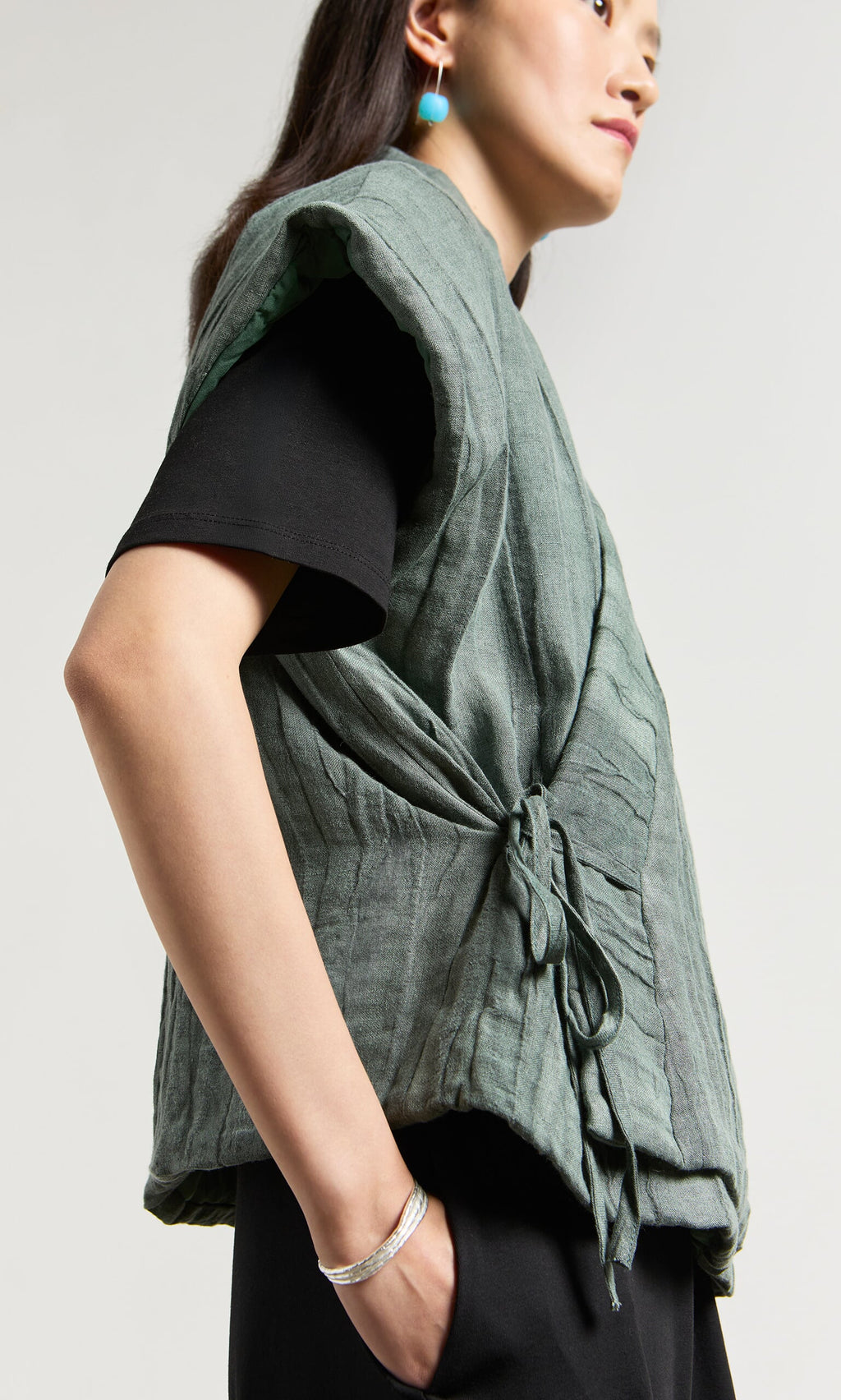 Plümo- Person wearing the Fawn Gilet with a textured woven finish over a black shirt and turquoise earrings, standing sideways.