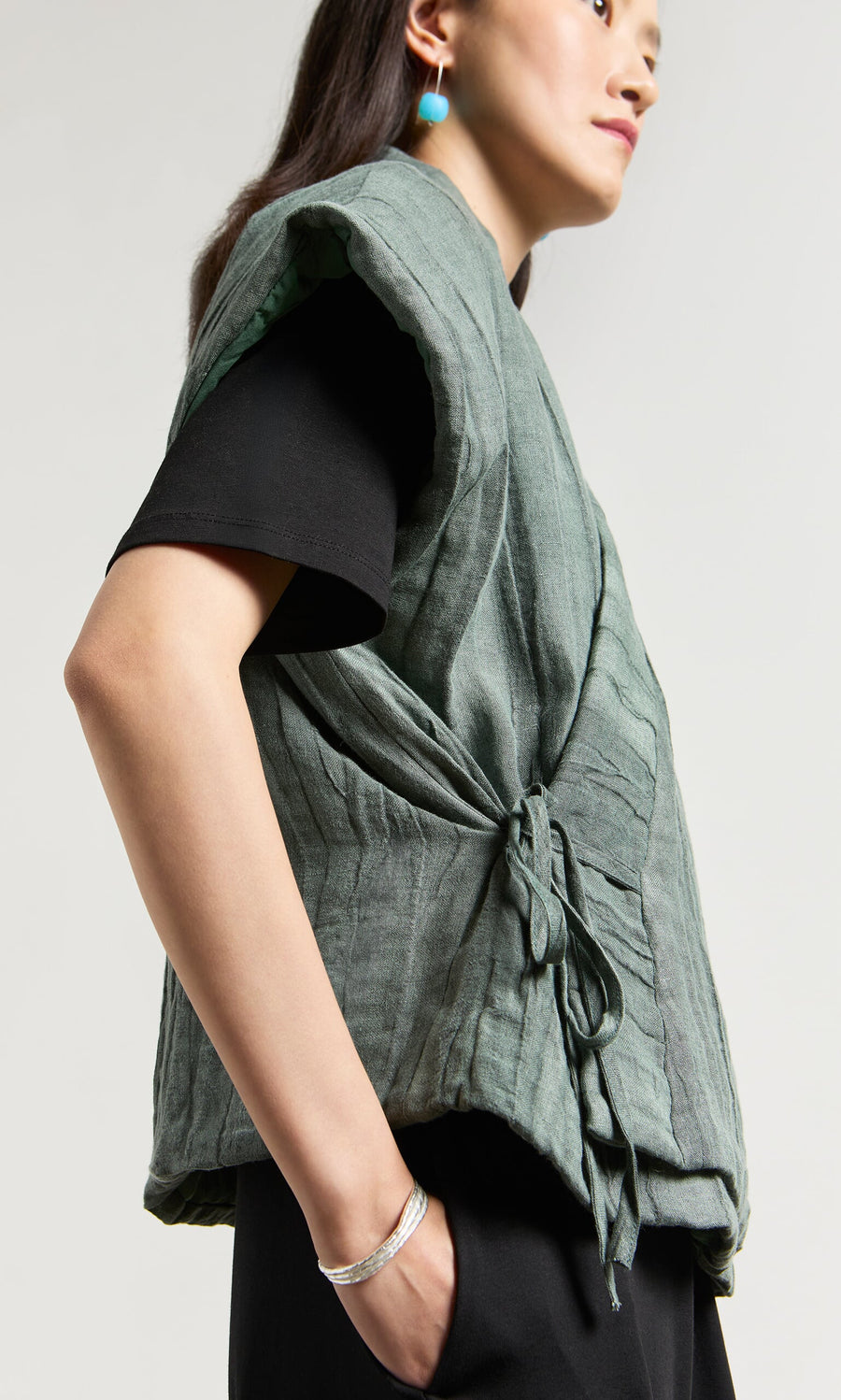 Plümo- Person wearing the Fawn Gilet with a textured woven finish over a black shirt and turquoise earrings, standing sideways.