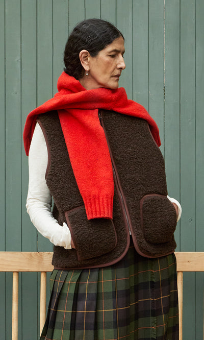 Plümo  Woman in a Feodor Gilet, red scarf, and plaid skirt stands against a green wall, gazing to the side.