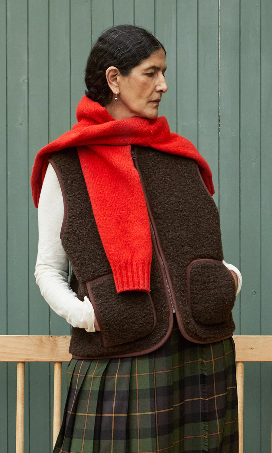 Plümo  Woman in a Feodor Gilet, red scarf, and plaid skirt stands against a green wall, gazing to the side.