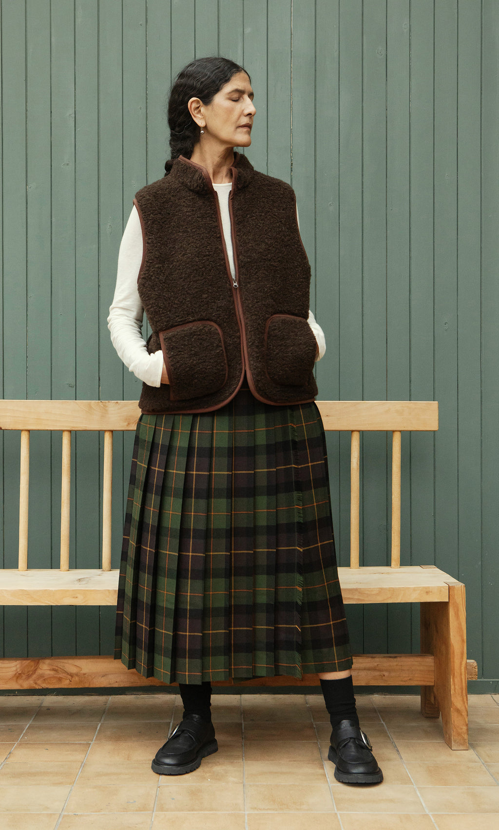 Plümo  Woman in the Feodor Gilet and plaid skirt stands by a wooden bench, looking left against a green paneled wall.