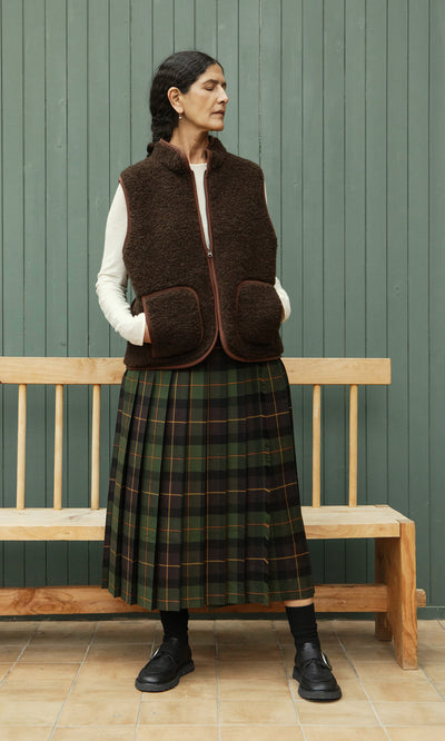 Plümo  A woman in a brown vest and Derry Kilt stands before a wooden bench and green paneled wall.