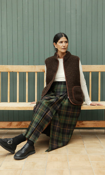 Plümo  Woman in a Derry Kilt and cream top sits on a wooden bench against a green paneled wall.