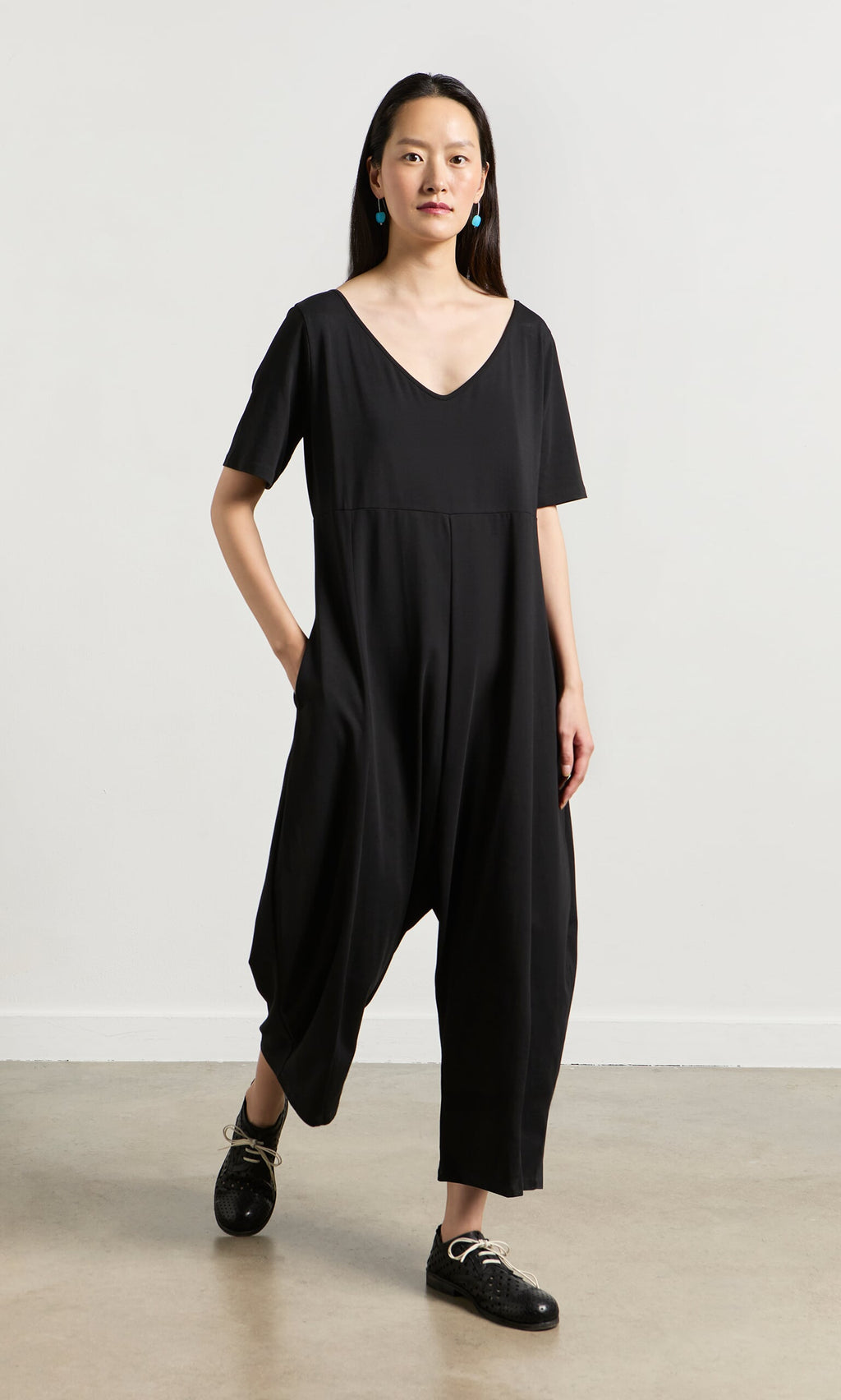 Plümo- Woman in Fenzi Jumpsuit—an oversized black, short-sleeve style—paired with black shoes and blue earrings.