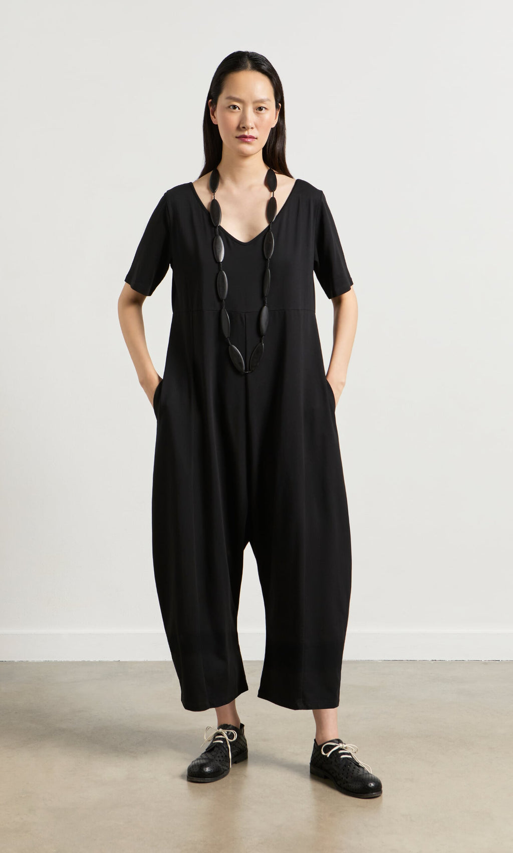 Plümo- Woman wearing the Fenzi Jumpsuit, an oversized black V-neck style, with chunky necklace and black shoes, hands in pockets.