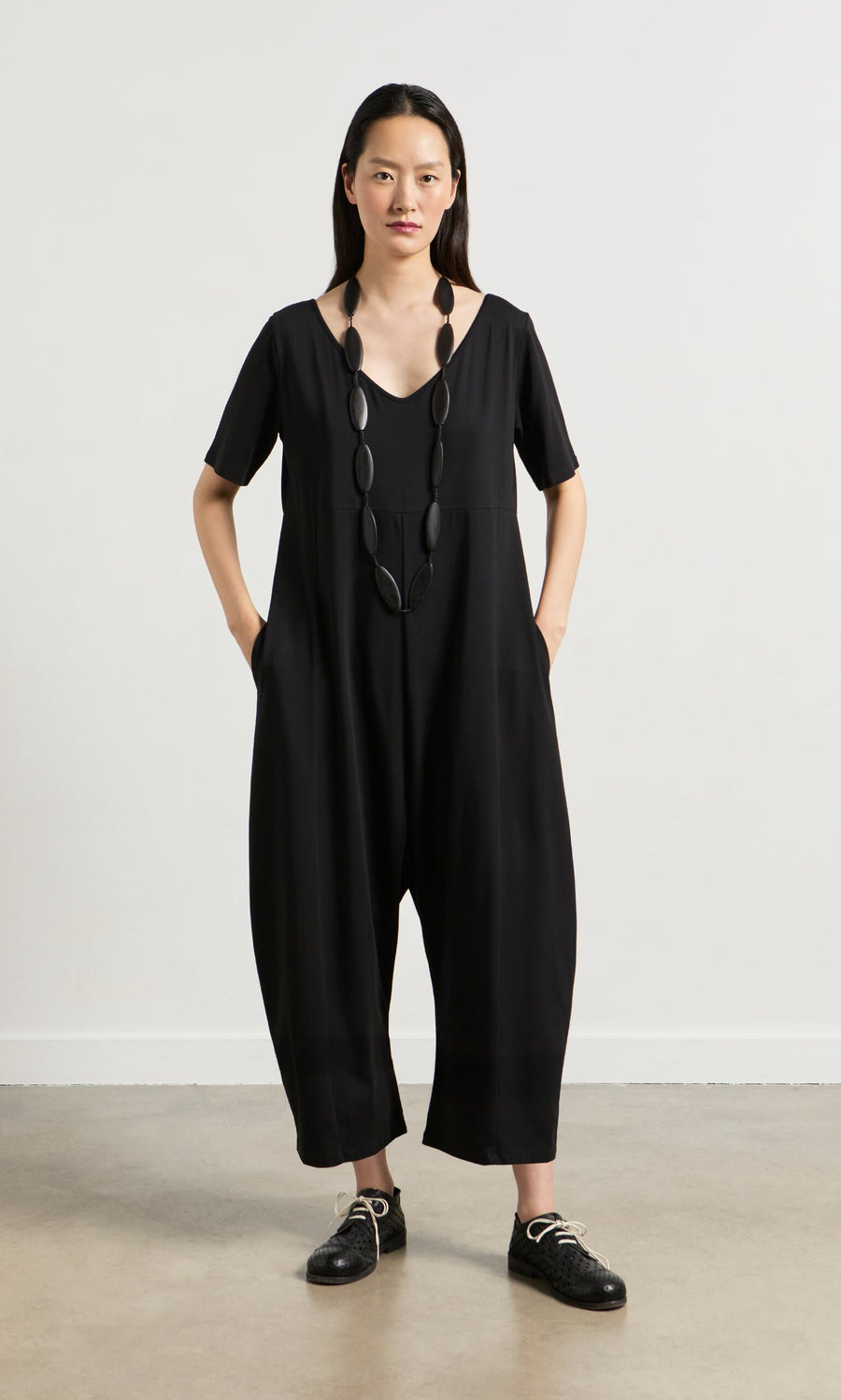 Plümo- Woman wearing the Fenzi Jumpsuit, an oversized black V-neck style, with chunky necklace and black shoes, hands in pockets.