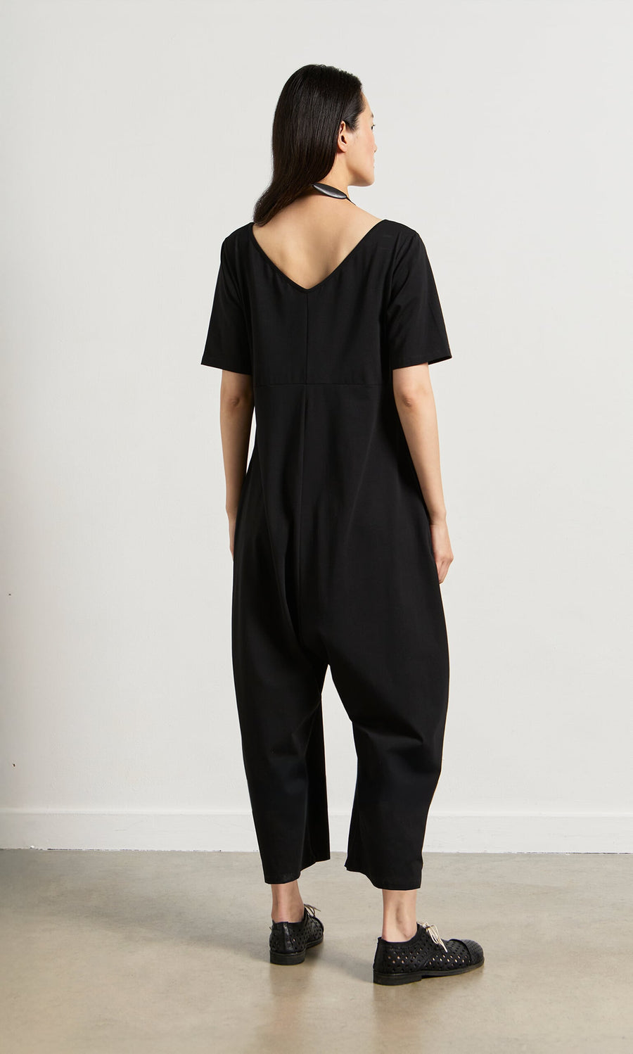 Plümo- Woman indoors, facing away, wearing the Fenzi Jumpsuit—an oversized black V-neck with V-cut back and black shoes.