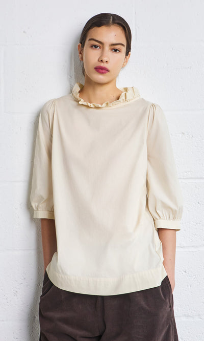 Plümo- Woman wears the Flint Blouse—ivory cotton with pie crust frill and puff sleeves—against a white wall.