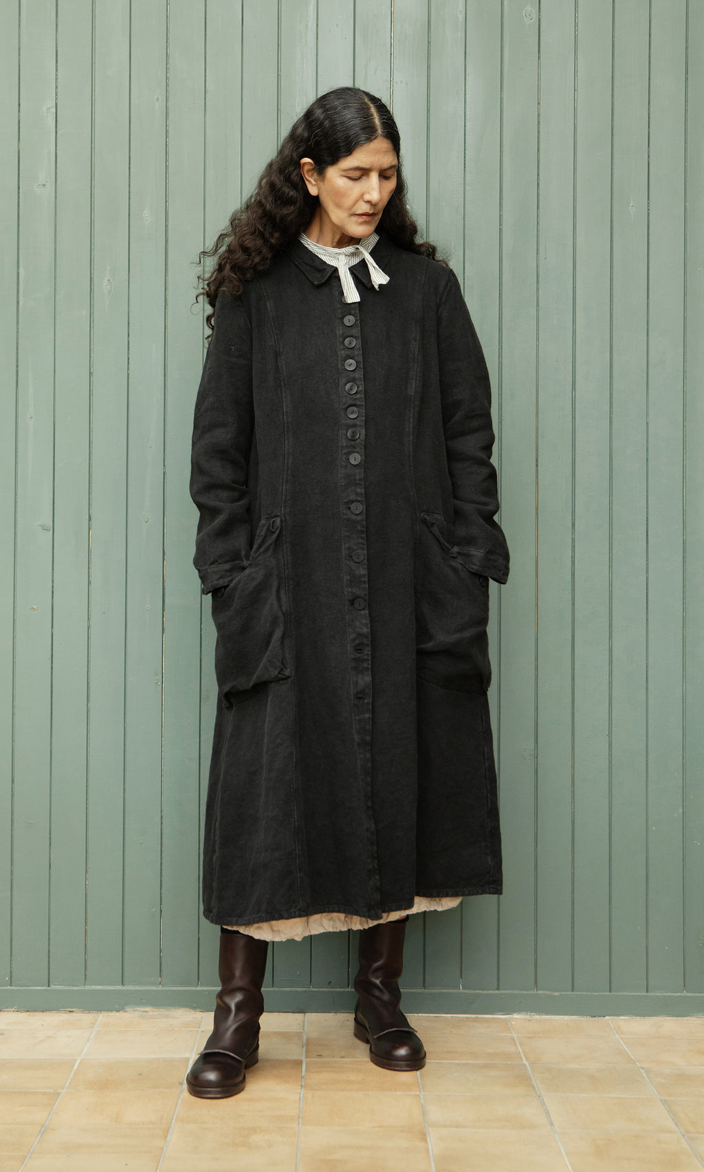 Plümo  Woman with long dark hair wears the relaxed fit Fowey Coat in black linen and brown boots, standing by a green wall.
