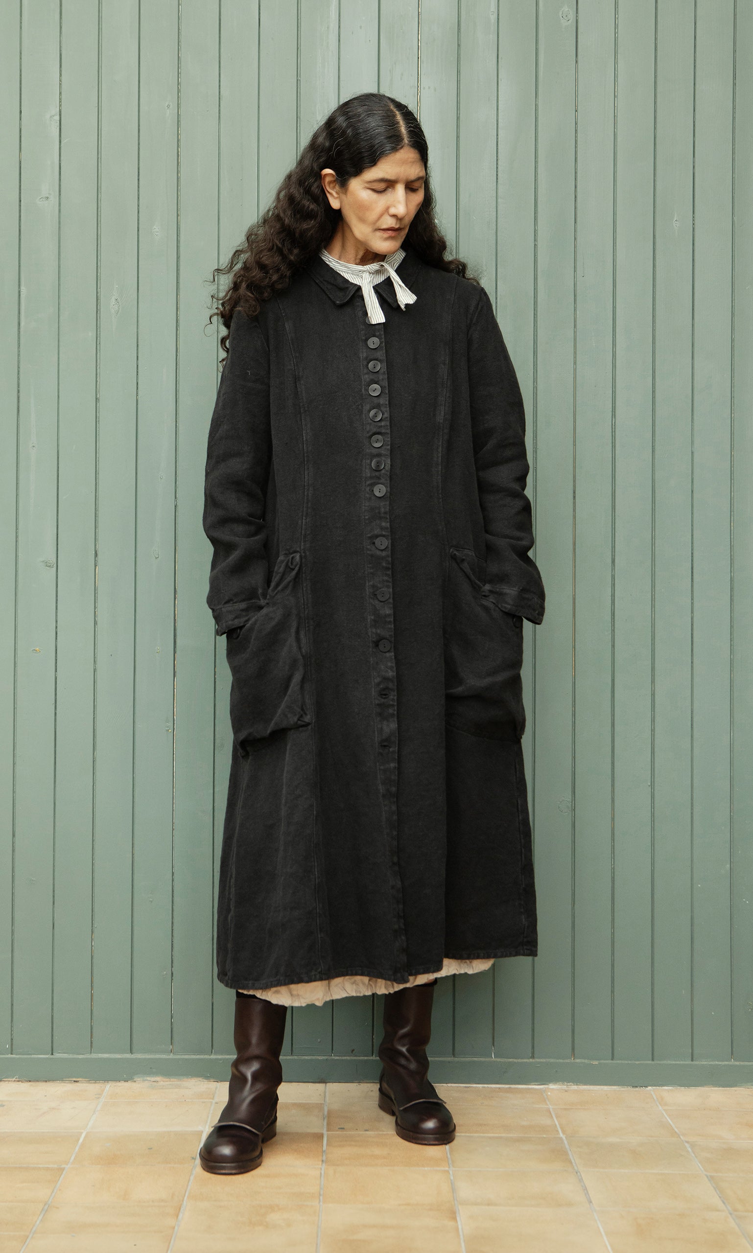 Plümo  Woman with long dark hair wears the relaxed fit Fowey Coat in black linen and brown boots, standing by a green wall.