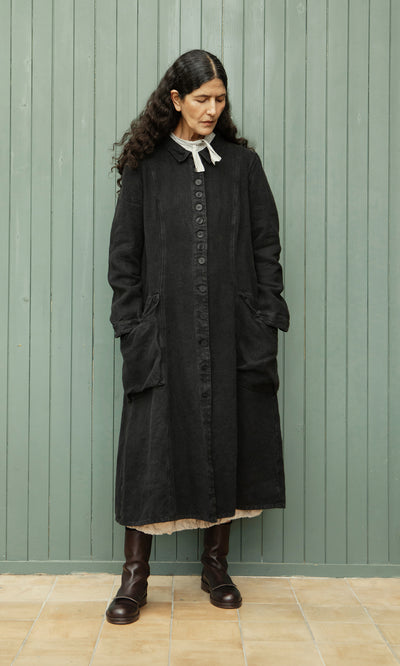 Plümo  Woman with long dark hair wears the relaxed fit Fowey Coat in black linen and brown boots, standing by a green wall.