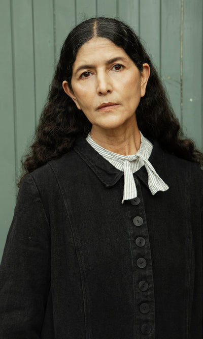 Plümo  Woman with long curly hair in the Fowey Coat, relaxed fit black linen, and striped bow tie by a green paneled wall.