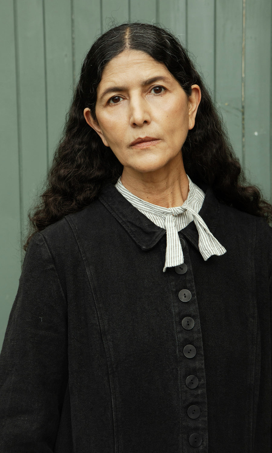 Plümo  Woman with long curly hair in the Fowey Coat, relaxed fit black linen, and striped bow tie by a green paneled wall.