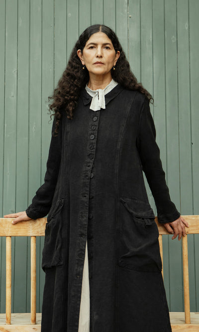 Plümo  Woman with long curly hair models the Fowey Coat in black linen, standing by a green wall with hands on a bench.