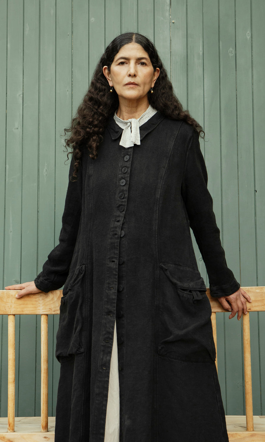 Plümo  Woman with long curly hair models the Fowey Coat in black linen, standing by a green wall with hands on a bench.