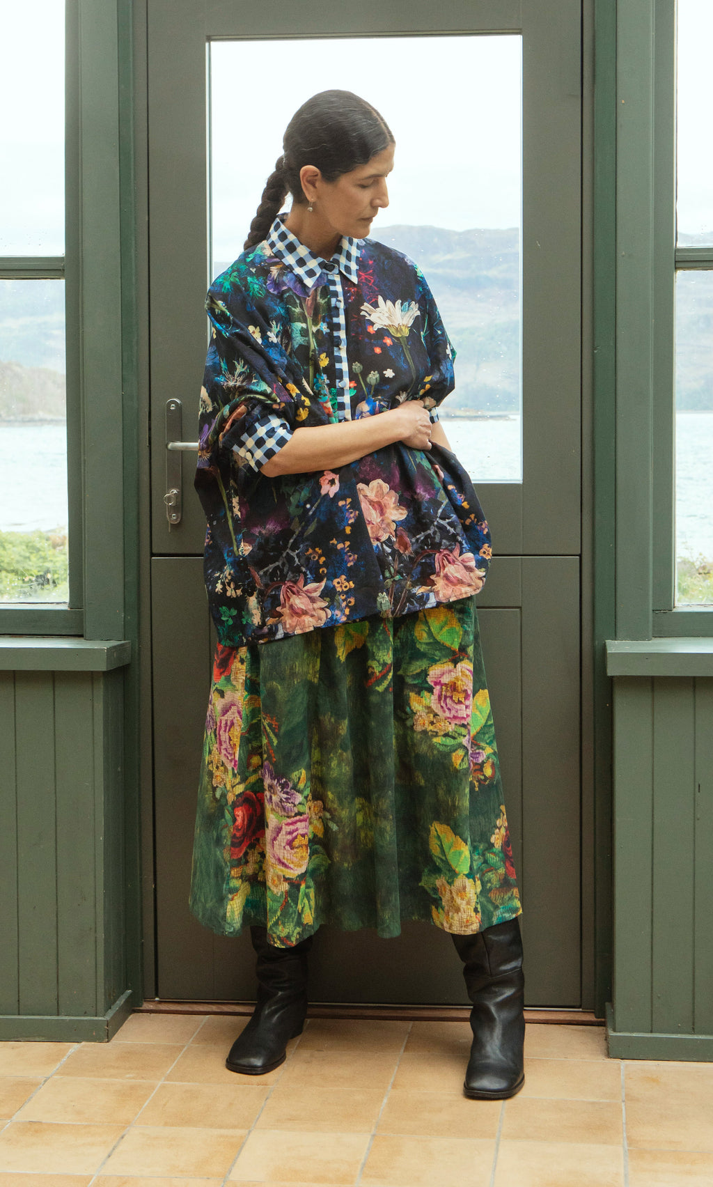 Plümo  Woman wears Foye Flower Shirt with layered florals, gingham collar, black boots; stands indoors by green door, looking down.