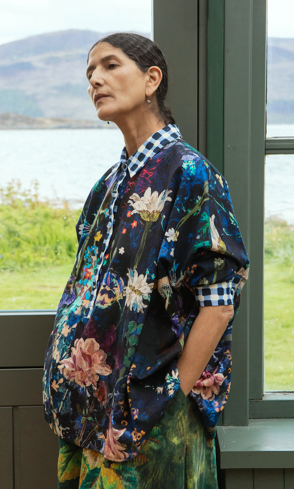 Plümo  Woman in the Foye Flower Shirt stands by a window, looking thoughtful with her hands in her pockets.