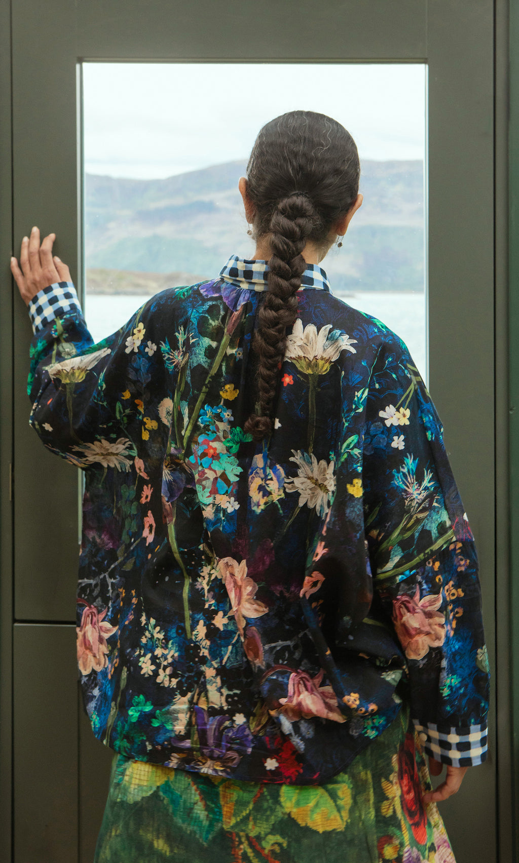 Plümo  Someone in a Foye Flower Shirt with a braid stands facing a window, mountains and water outside.