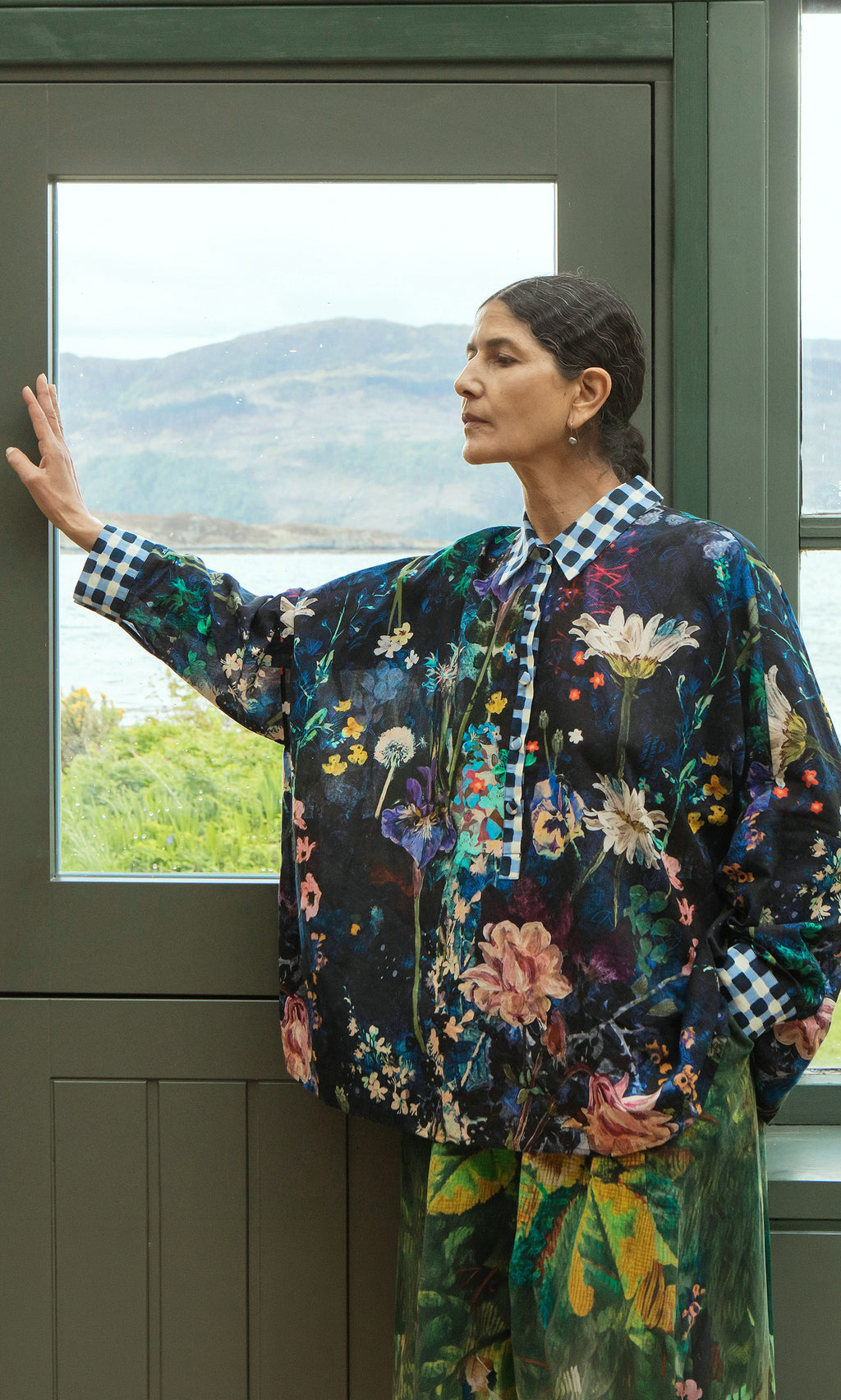 Plümo  Woman in Foye Flower Shirt with gingham collar stands by window, gazing outside with one hand on the frame.