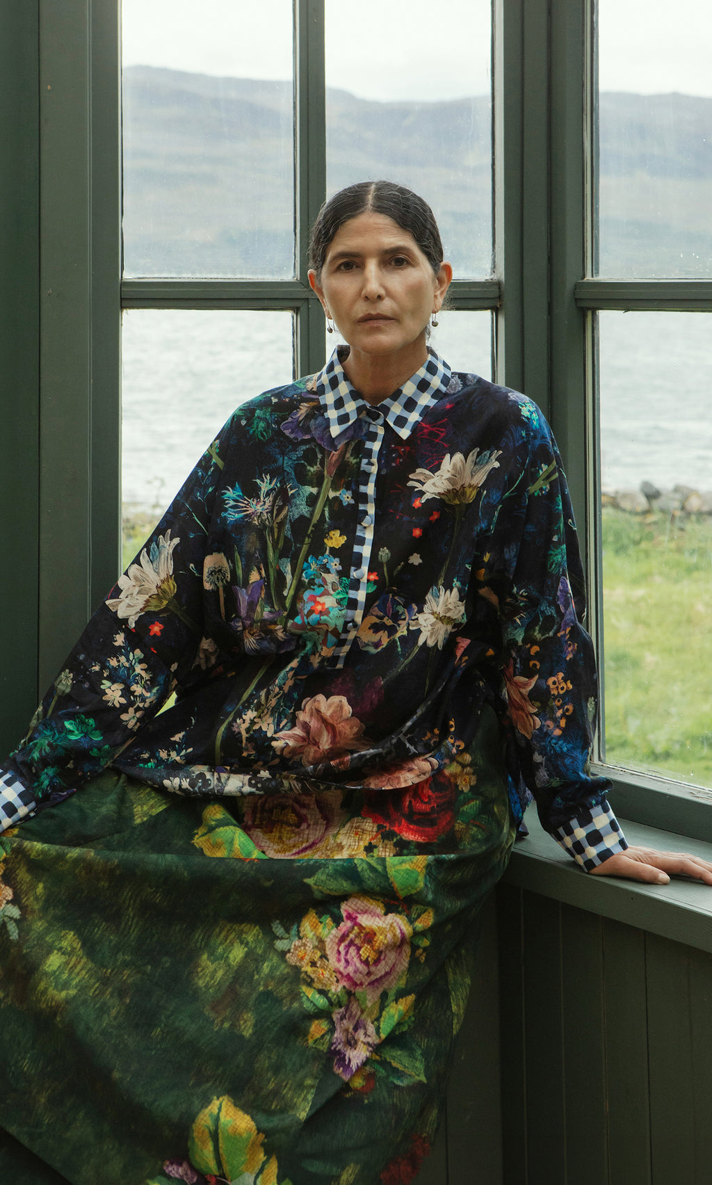 Plümo  A woman in a Foye Flower Shirt sits on a windowsill, facing the camera, with a lake and hills in the background.