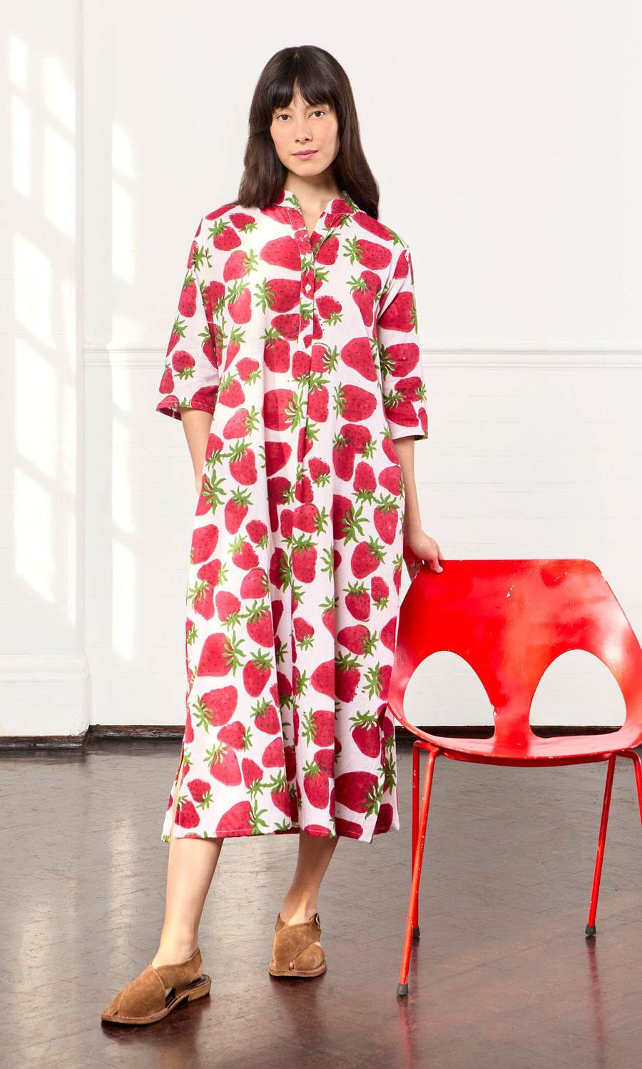 Plümo- Woman in the Fresa Dress, a cotton voile shirt dress with strawberry block print, stands by a red chair in sunlight.