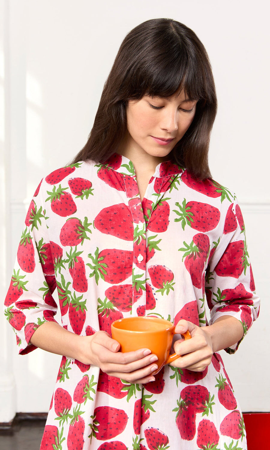 Plümo- Woman in the Fresa Dress holds an orange mug, looking down in a bright room.