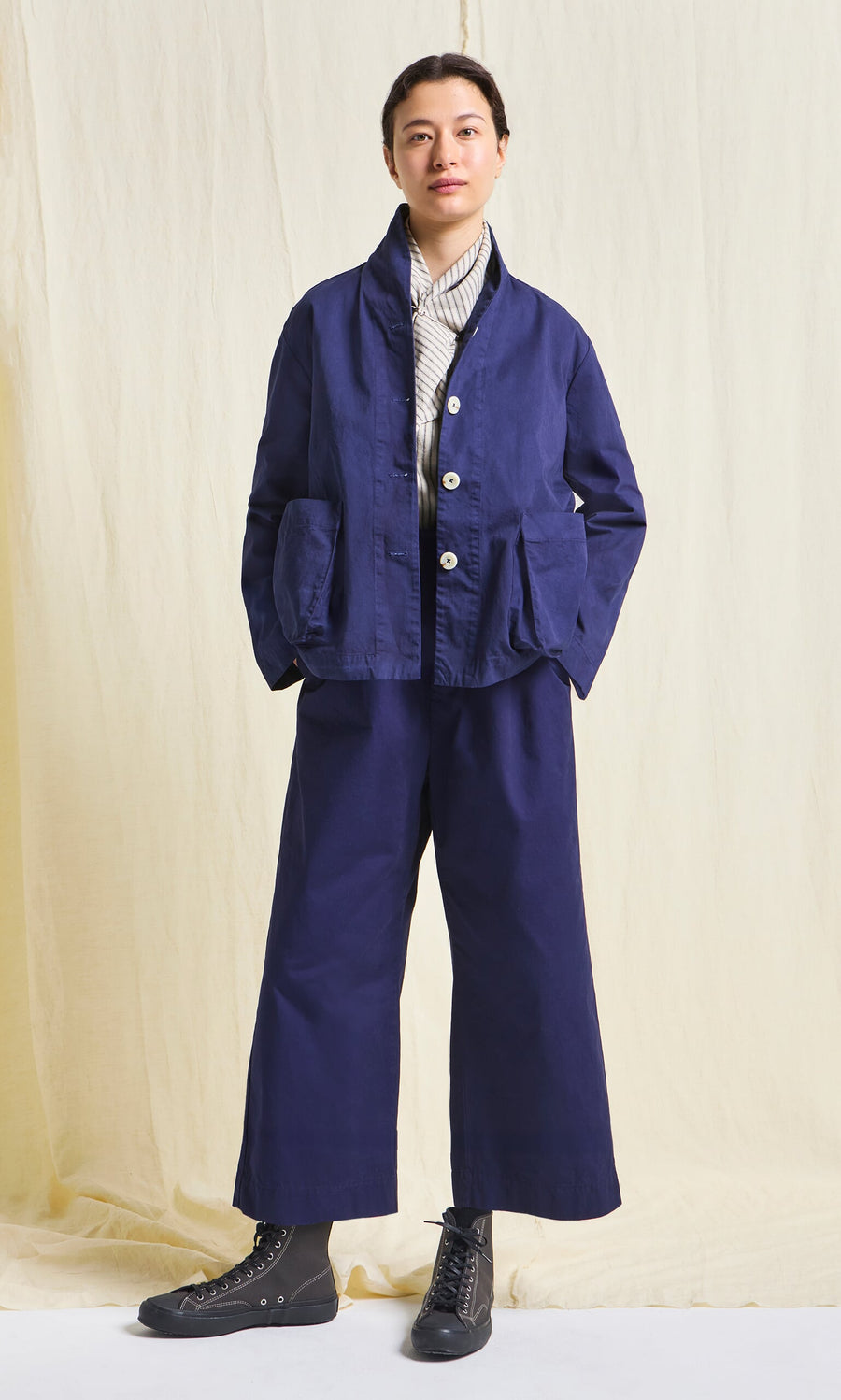 Plümo- Person in the Gardener's Jacket - Navy, wide-leg pants, striped shirt, black boots, against a beige backdrop.