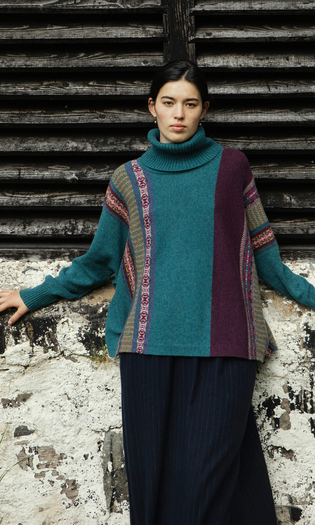Plümo  Woman in a Greba Rollneck teal and purple fairisle sweater stands by a weathered wall with dark wooden slats.