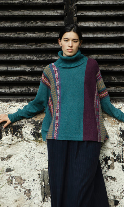 Plümo  Woman in a Greba Rollneck teal and purple fairisle sweater stands by a weathered wall with dark wooden slats.