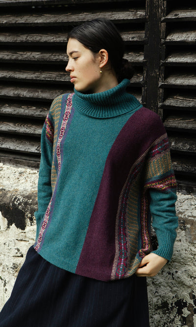 Plümo  Woman in a teal and purple Greba Rollneck stands by a weathered wall and wooden shutters, gazing to the left.