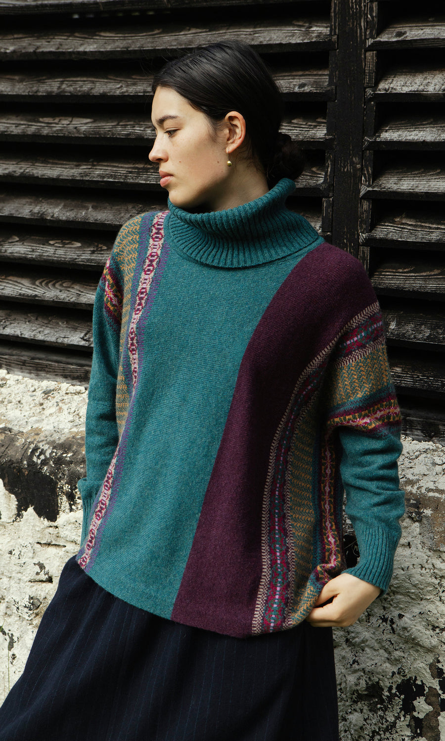 Plümo  Woman in a teal and purple Greba Rollneck stands by a weathered wall and wooden shutters, gazing to the left.