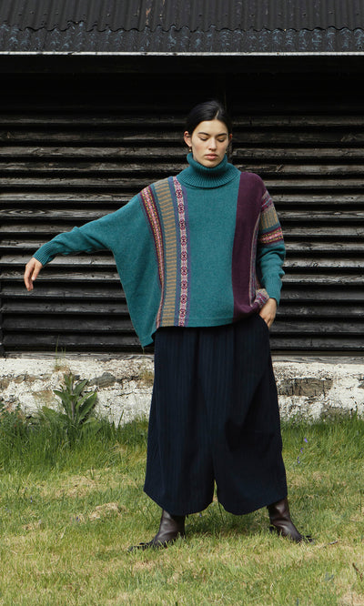 Plümo  Woman in the Greba Rollneck sweater and wide black pants stands on grass by a rustic wooden building.