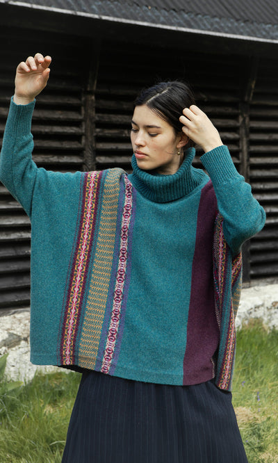 Plümo  Person in a Greba Rollneck stands outdoors, one arm raised and hand touching hair by a wooden wall.