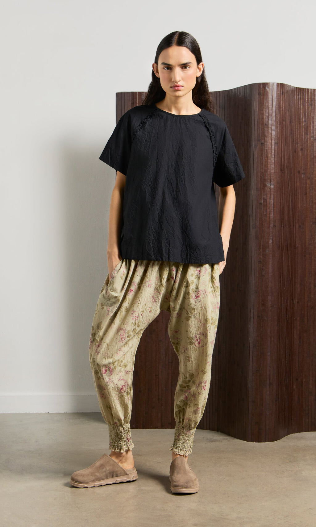 Plümo- Woman in a black short-sleeve top and Green Floral Pantaloons stands in front of a brown folding screen.