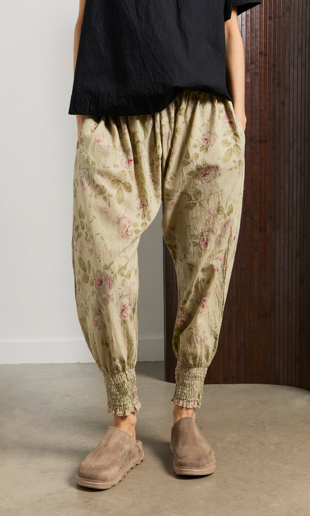 Plümo- Person wears Green Floral Pantaloons, a black top, and taupe slip-on shoes indoors with hands in pockets.