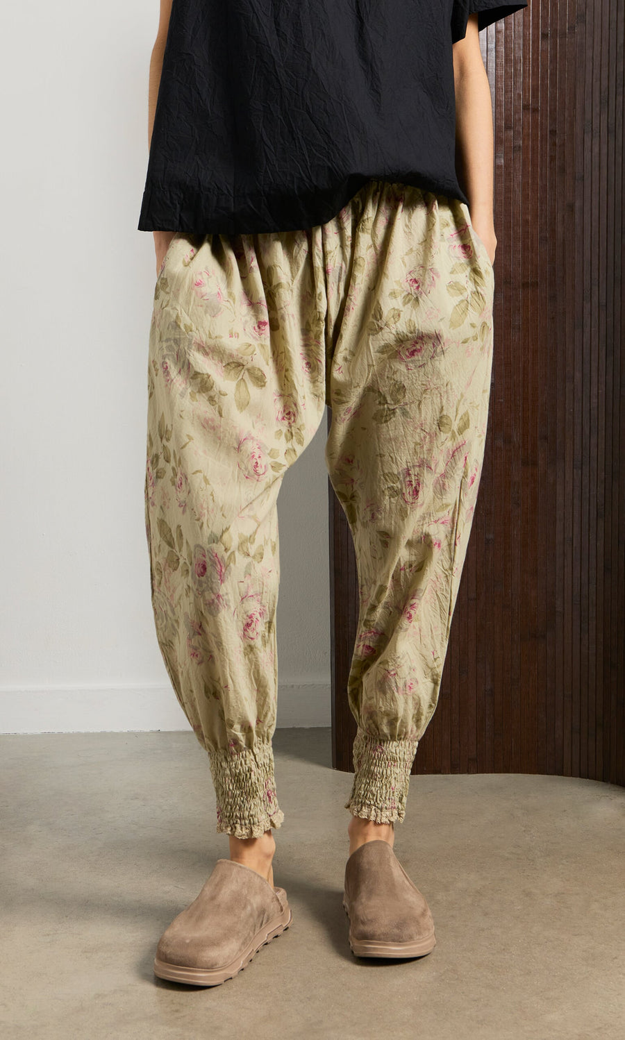 Plümo- Person wears Green Floral Pantaloons, a black top, and taupe slip-on shoes indoors with hands in pockets.
