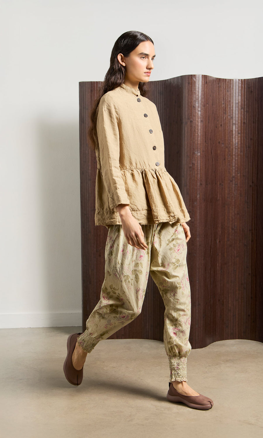 Plümo- Person wearing Green Floral Pantaloons and beige top walking indoors, brown folding screen in background.