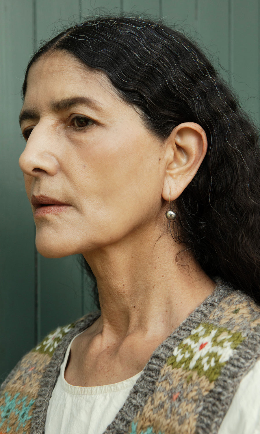 Plümo  Woman with long dark hair wears Greta Earrings - Silver and a patterned vest, gazing left against a green backdrop.