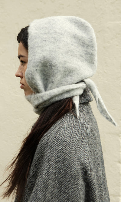 Plümo  Person in profile wears the Grey Bonnet, a Danish-designed woolen hood, standing against a plain light wall.