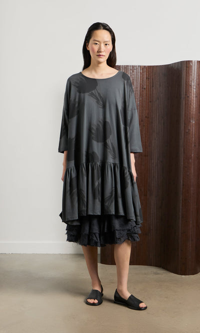 Plümo- Woman in a loose Grey Flower Blouse, black sandals, standing indoors by a brown folding screen.