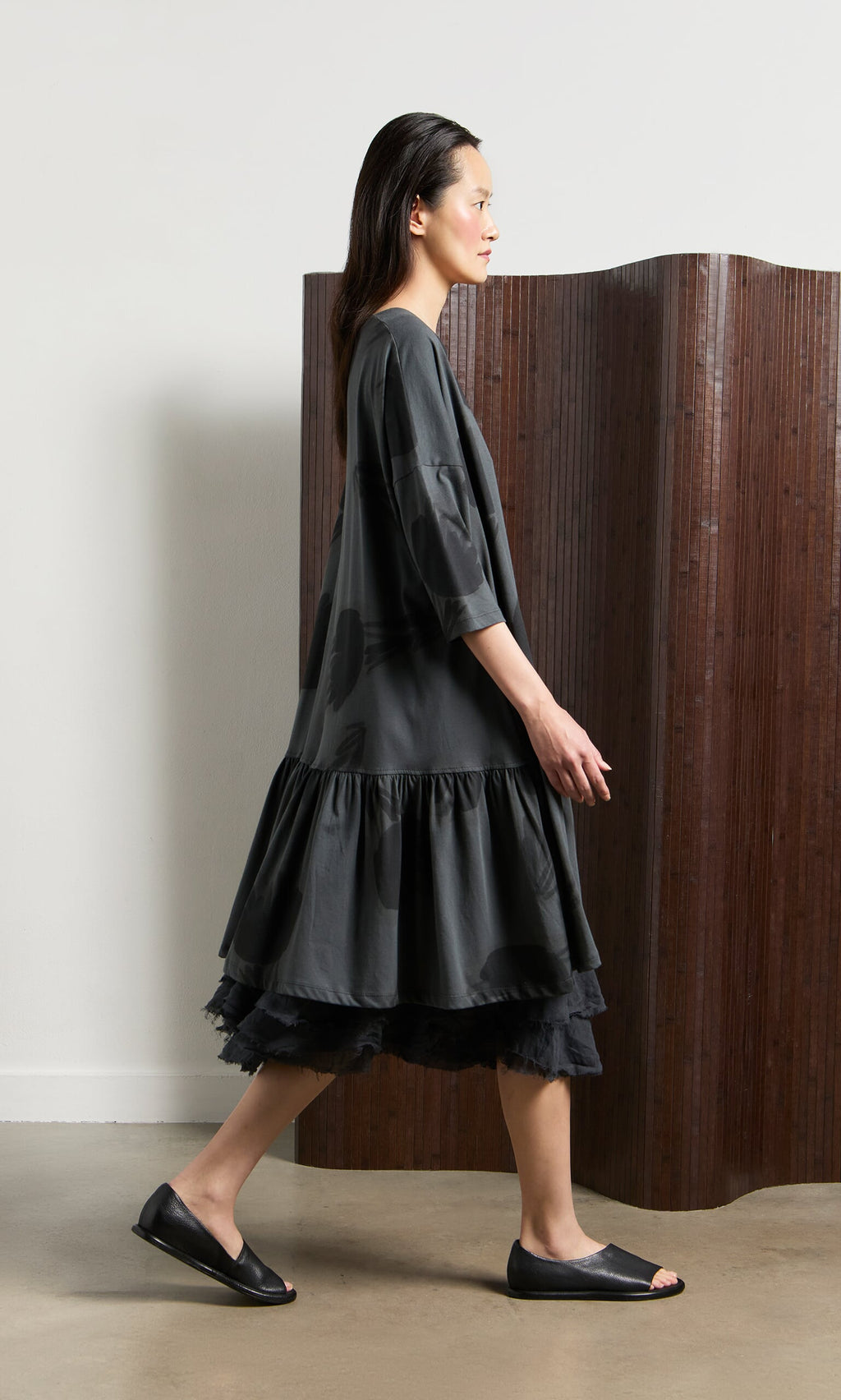Plümo- Woman in a loose dark dress, black flats, and Grey Flower Blouse walks indoors past a wooden room divider.