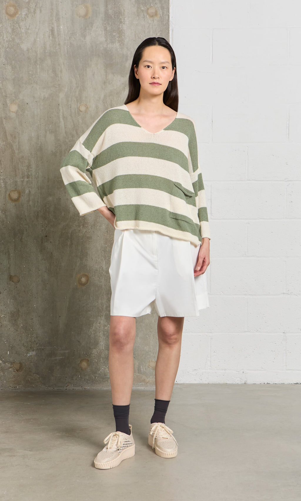 Plümo- Woman in a relaxed fit, sage and white Hagen Stripe Jumper, white shorts, and beige shoes stands by a concrete wall.