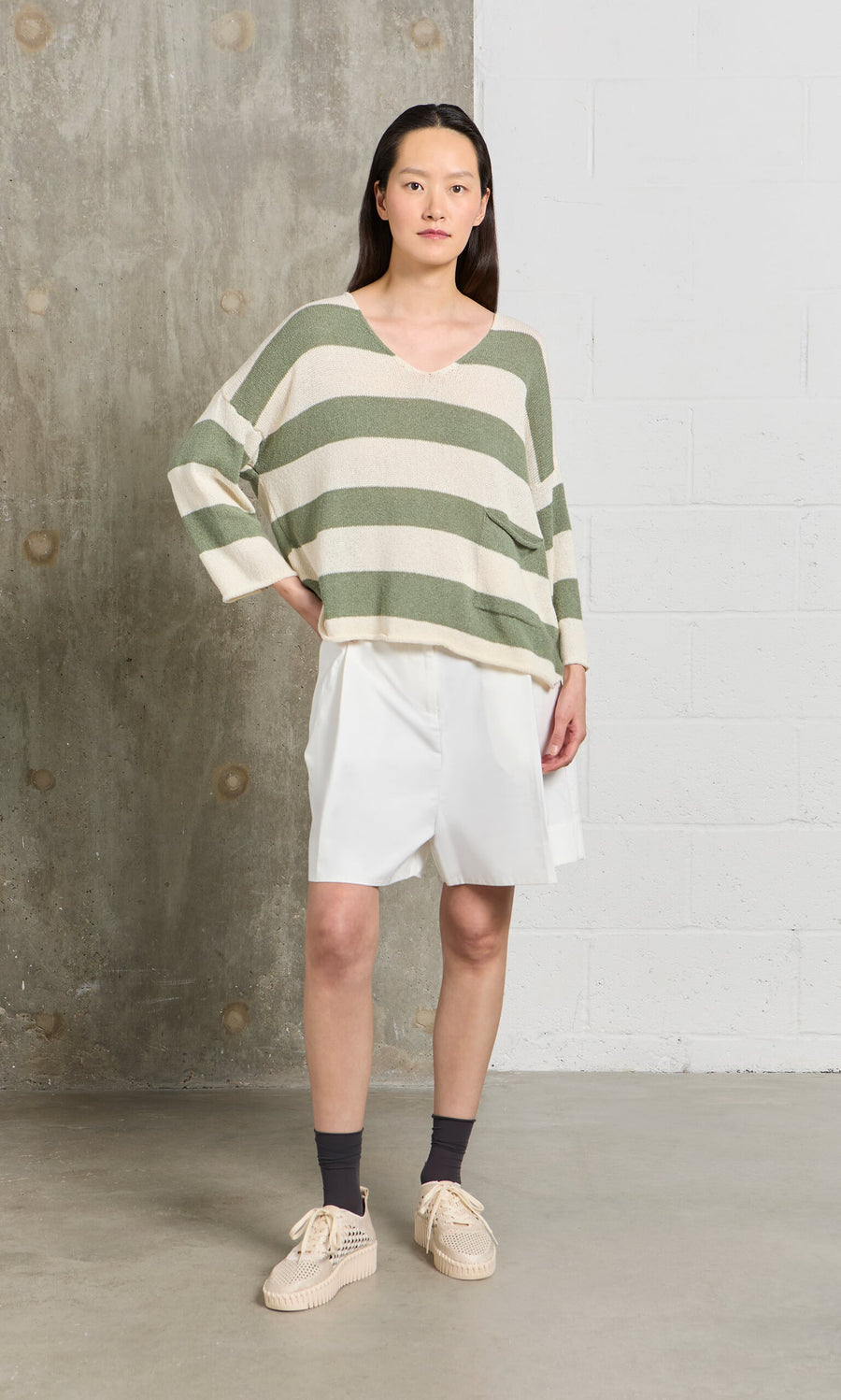 Plümo- Woman in a relaxed fit, sage and white Hagen Stripe Jumper, white shorts, and beige shoes stands by a concrete wall.
