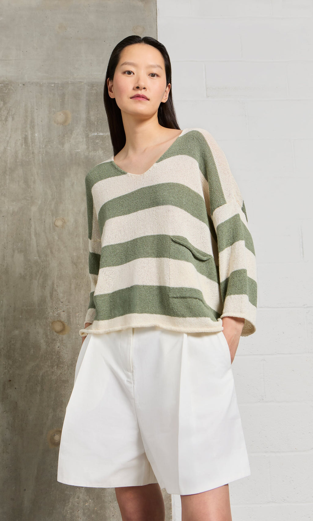 Plümo- Woman models the Hagen Stripe Jumper, a relaxed fit sage and white V-neck sweater, with white shorts.